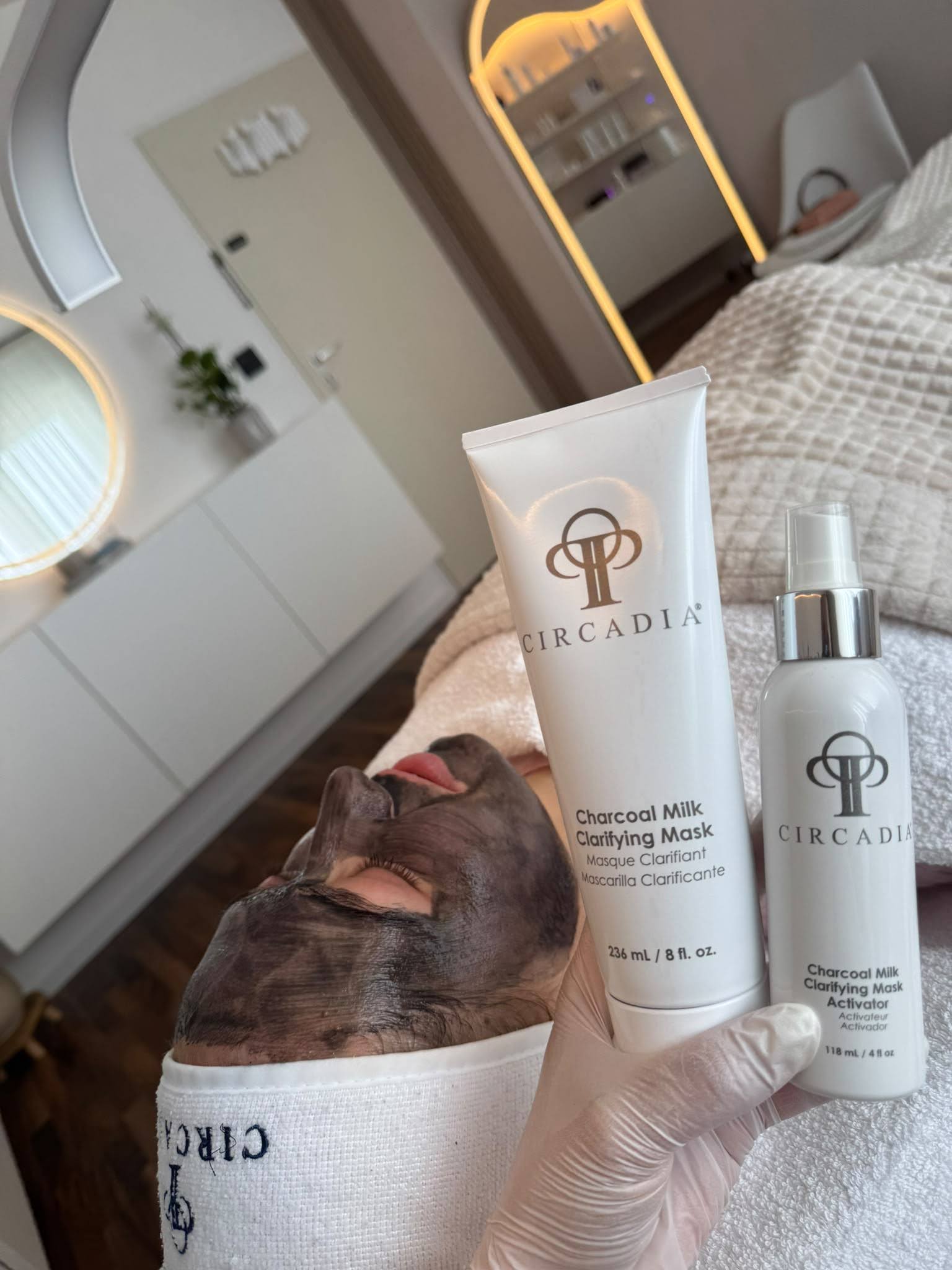 Behandschuhte Hand hält die Circadia Charcoal Milk Clarifying Mask und den zugehörigen Aktivator, im Hintergrund eine Kundin mit Aktivkohlemaske