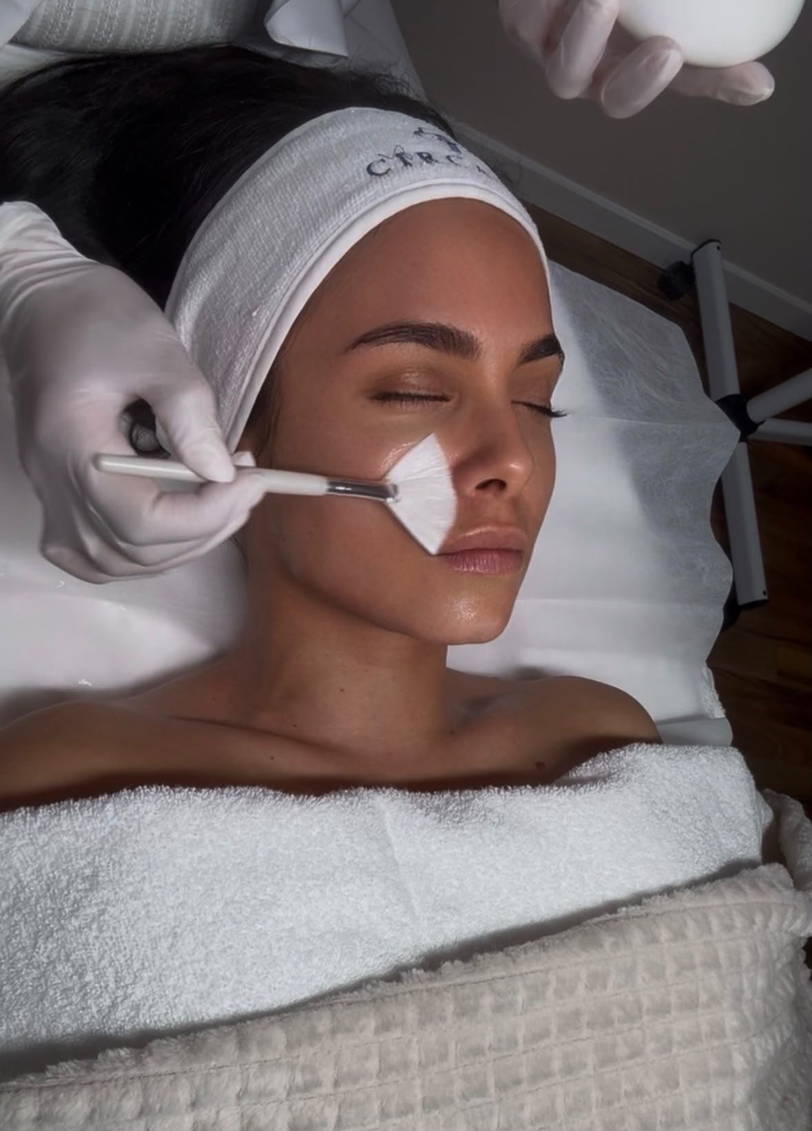Behandschuhte Hände tragen mit einem Fächerpinsel eine weiße Wirkstoffmaske auf die Wange einer liegenden Kundin auf, die ein Circadia-Stirnband trägt