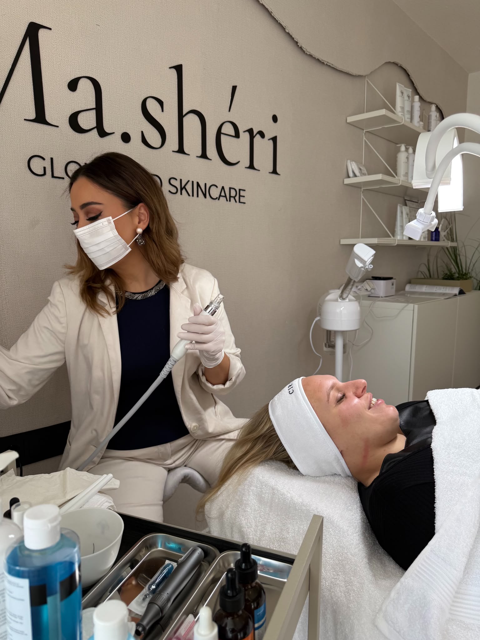 Kosmetikerin mit Mundschutz und Handschuhen hält ein Behandlungshandstück, im Hintergrund der Wandschriftzug Ma.shéri Glow and Skincare und ein Regal mit Pflegeprodukten