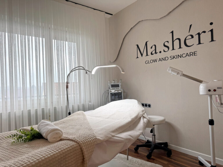Frisch bezogene Behandlungsliege mit gefalteten Handtüchern und einem Eukalyptuszweig, darüber eine Lupenleuchte, im Hintergrund die Wand mit dem Schriftzug Ma.shéri Glow and Skincare