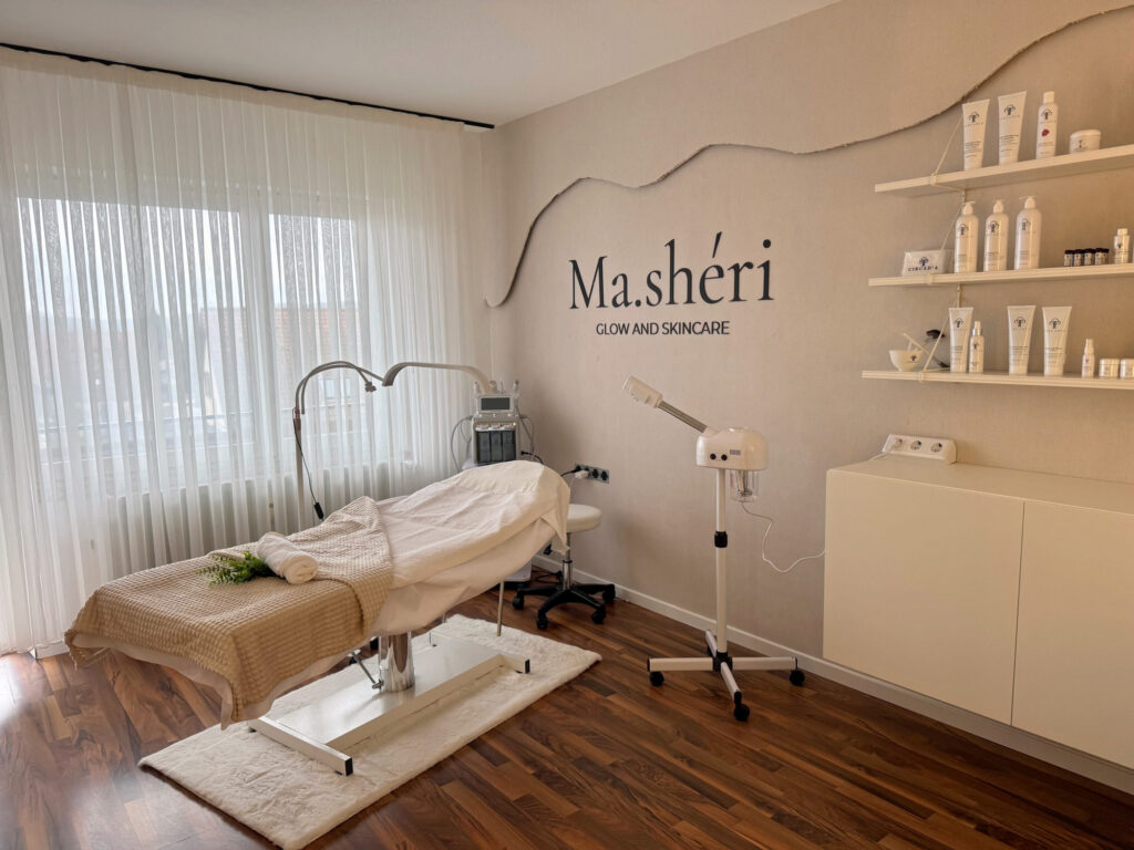 Heller Behandlungsraum mit gepolsterter Liege vor einem bodentiefen Fenster mit Vorhang, daneben Lupenleuchte und Bedampfer, an der Wand der Schriftzug Ma.shéri Glow and Skincare und Regale mit Pflegeprodukten