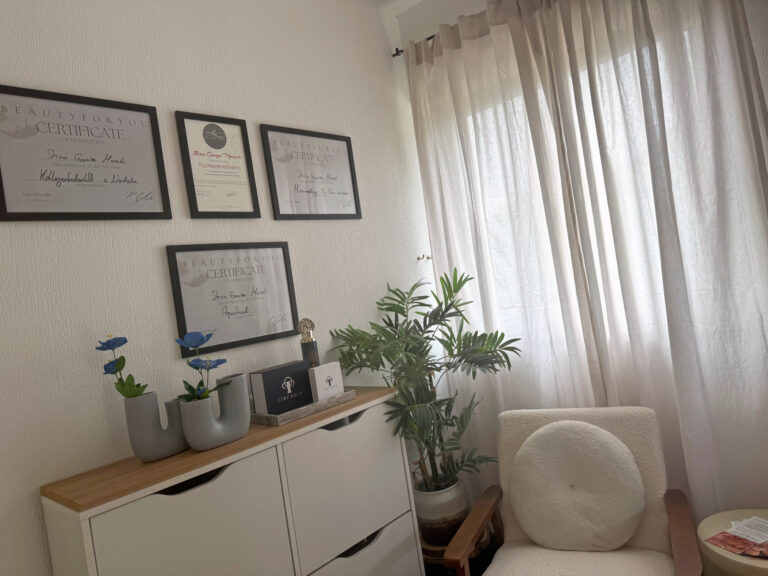Ruhige Sitzecke mit einem weißen Bouclé-Sessel am Fenster, daneben eine Zimmerpflanze und ein Sideboard, darüber vier gerahmte Fortbildungszertifikate